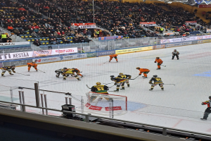Eine Gruppe von Menschen, die Hockey auf einem Eisring mit einem Netz in der Mitte spielen, umgeben von einem Zaun, mit Zuschauern im Hintergrund und Bannern mit Text, unter Stadionbeleuchtung.