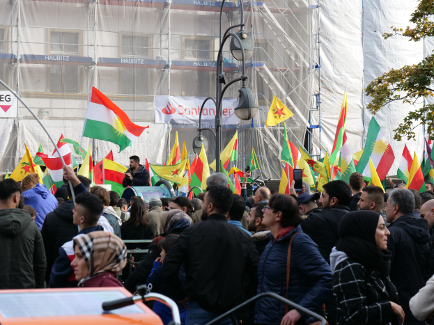 Eine große Gruppe von Menschen mit Fahnen vor einem Gebäude versammelt, eine Person in der Mitte mit einem Mikrofon, ein Fahrzeug unten, ein Baum rechts und Stangen, Lichter und Banner mit Text im Hintergrund, wahrscheinlich eine kurdische Demonstration in Istanbul.