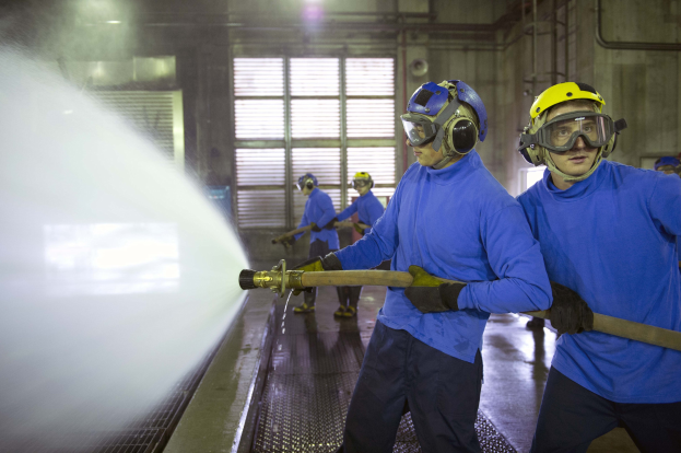 Eine Gruppe von Männern in blauen Hemden und gelben Helmen arbeitet an einer Maschine in einer Fabrikumgebung, wobei einer einen Schlauch hält und Wasser auf den Boden sprüht.