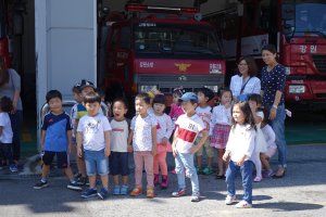Eine Gruppe von Kindern vor einem Feuerwehrauto an einer Feuerwache stehend, einige mit M√∞tzen, mit weiteren Feuerwehrautos im Hintergrund.