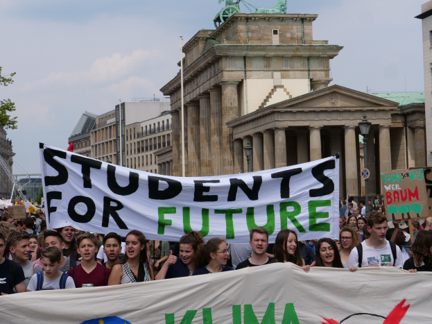 Gruppe von Schülern marschiert in Berlin mit einem bunten "Students for Future"-Schild vor Gebäuden, Bäumen und Himmel.