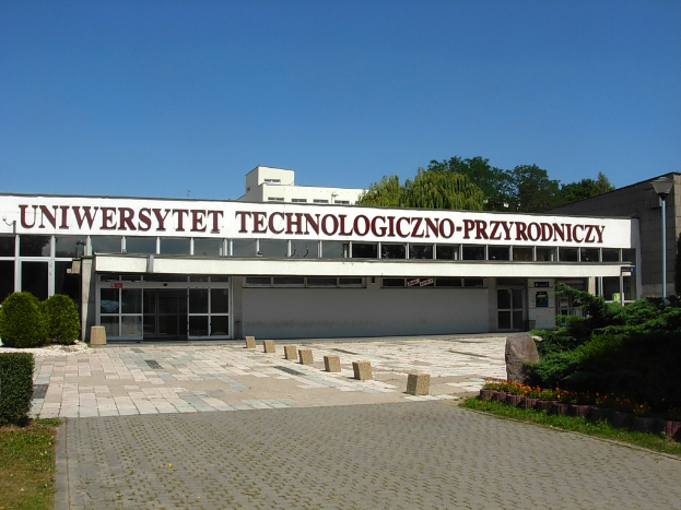 Die Technische Universität Prag, ein großes Gebäude mit mehreren Fenstern und Türen, umgeben von Grünflächen, Bäumen und einem Straßenpfahl, unter einem bewölkten Himmel.