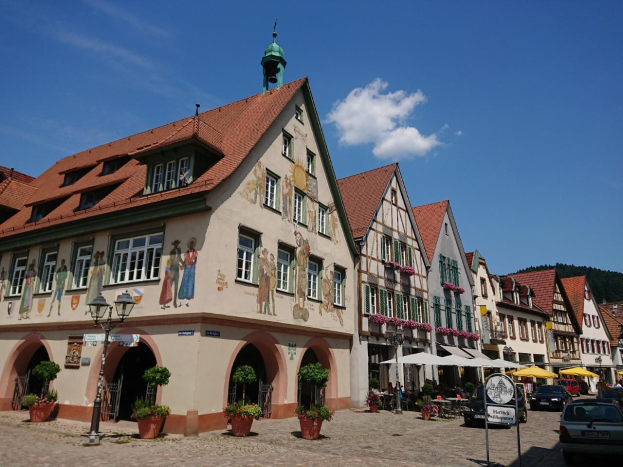 Ein Kopfsteinpflasterweg in Rothenburg ob der Tauber, Deutschland, gesäumt von Gebäuden, Laternen, Blumentöpfen, Schirmen und Fahrzeugen, mit Bäumen und einem bewölkten Himmel im Hintergrund.