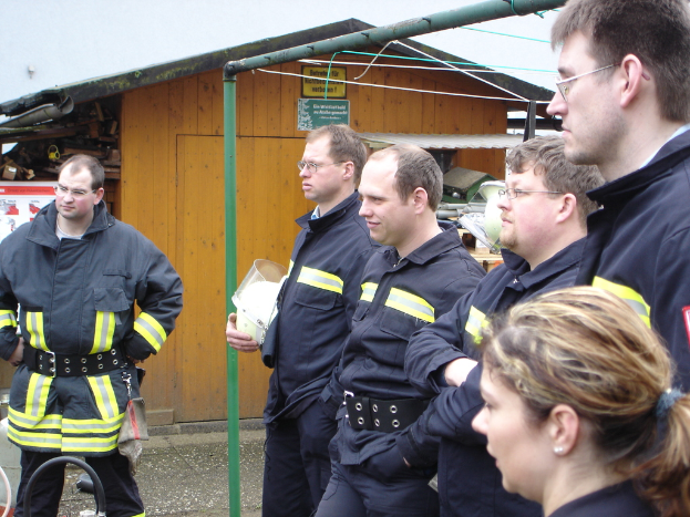 Eine Gruppe von Männern in blauen Feuerwehreinheitsuniformen steht nach links gewandt, mit einem kleinen hölzernen Schuppen und Rohrrahmen im Hintergrund.