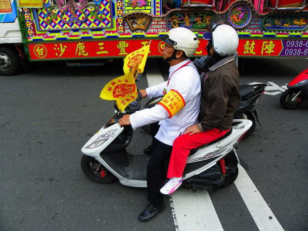 Zwei Personen mit Helmen fahren auf einem Motorroller, eine hält eine gelbe Fahne, mit einem Fahrzeug im Hintergrund.