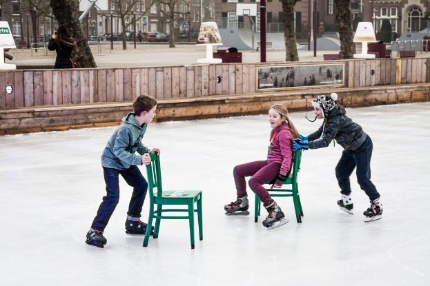 Kinder beim Skifahren in einem Spielplatz mit drei Kindern in der Mitte, zwei Stühlen, Gebäuden, Bäumen, Bänken, Pfosten und einem Basketballfeld im Hintergrund.
