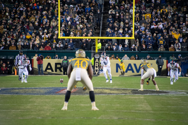 Fußballspieler mit Helmen und Uniformen konkurrieren auf einem Feld mit Zuschauern in den Rängen und Stadionwerbung im Hintergrund.