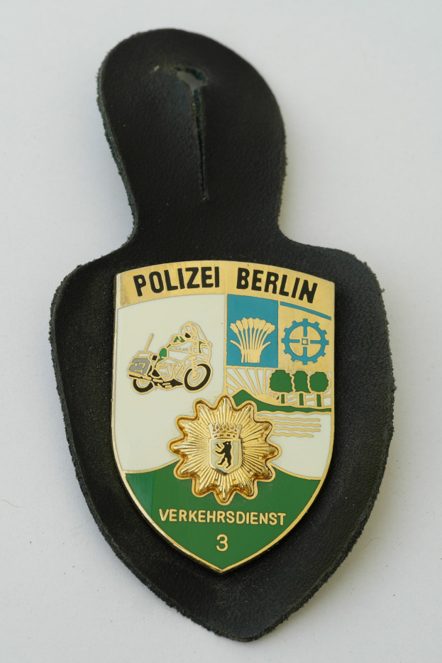 Ein schildförmiges Polizeibadgebnis mit der Aufschrift 'Polizei Berlin' auf einem blauen Hintergrund, einem weißen Rand, einem weißen zentralen Stern und schwarzer fetter Schrift auf einer Oberfläche.