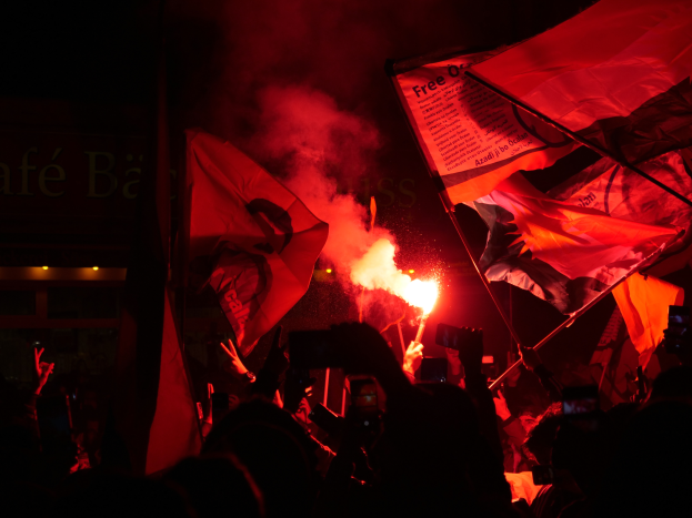 Eine Gruppe von Menschen mit Fahnen und Fackeln, beleuchtet vom Licht des Feuers, das sie halten, steht vor einem Gebäude mit Text darauf, was auf einen Protest gegen Regierungssparmaßnahmen hindeutet.