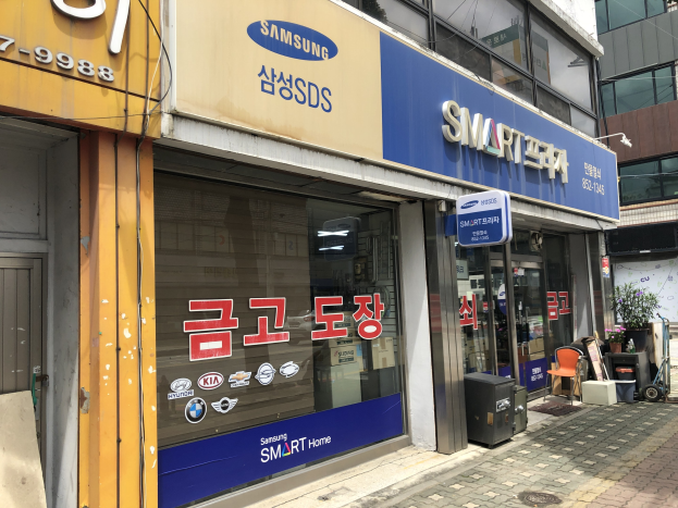 Außenansicht eines Gebäudes mit einer 'Samsung Smart Home'-Plakette, umgeben von Glaswänden mit Namensschildern, mit Mülltonnen, Stühlen, Pflanzen und einem sichtbaren Himmel im Hintergrund.