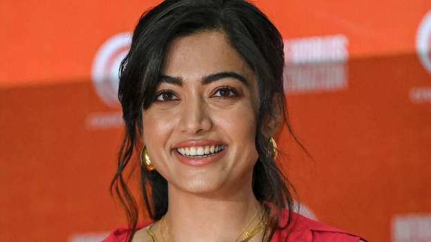Eine Frau in einem roten Kleid mit einem eleganten Dutt, schwarzen High Heels und dezentem Make-up lächelt in die Kamera vor einem gelben Banner mit der Aufschrift 'Die Besten Schönheits-Looks von Sonam Kapoor beim Cannes Film Festival.'