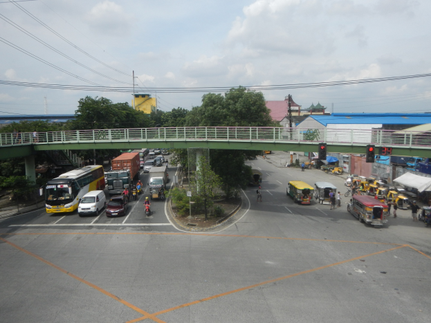 Eine belebte städtische Straße mit Autos, Bussen, Fußgängern auf einem Gehweg, einer Brücke mit Geländern, Verkehrsampeln, Bäumen, Gebäuden, Strommasten mit Drähten und einem bewölkten Himmel.