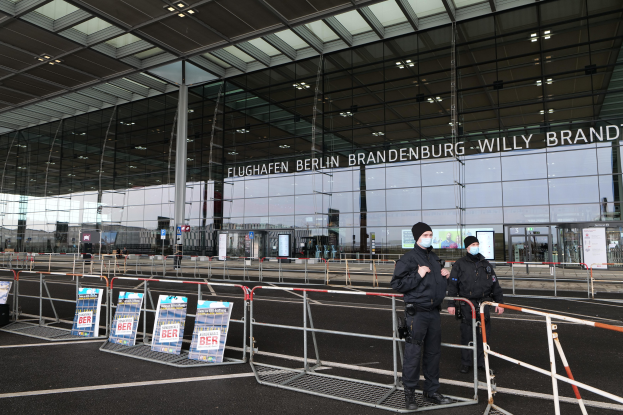 Zwei Polizisten in schwarzer Uniform stehen vor einem gläsernen Gebäude mit Text, mit Absperrungen und Schildern im Vordergrund.