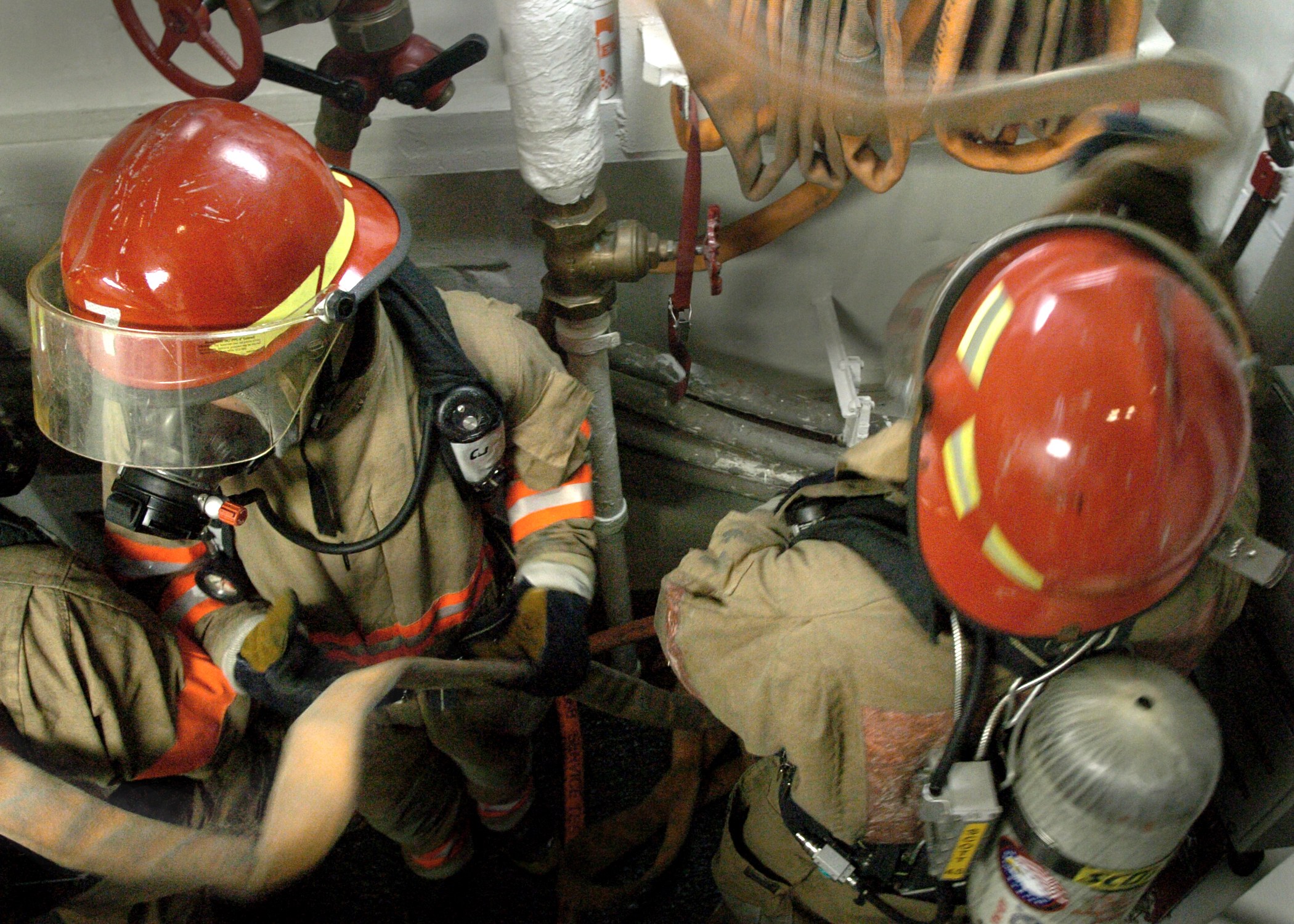 Feuerwehrleute in Schutzausrüstung bei der Arbeit an einem Hydranten mit Rohren und Ventilen im Hintergrund.