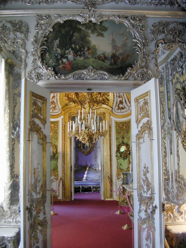 Kerzen an einem Kronleuchter, Türen mit Gemälden verziert und ein roter Teppich auf dem Boden.