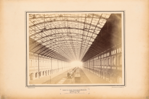 Ein altes Foto eines Bahnhofs mit einem Zug auf den Schienen, umgeben von Gebäuden mit Fenstern und einem Dach, das von Metallpfosten gestützt wird, mit Text am unteren Bildrand.