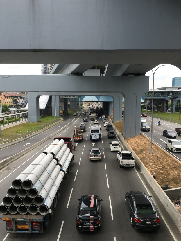 Eine belebte Autobahn unter einer Brücke mit zahlreichen Fahrzeugen, Pfählen, Lampen, Gras, Gebäuden, Bäumen und anderen Gegenständen im Hintergrund, unter einem Himmel.