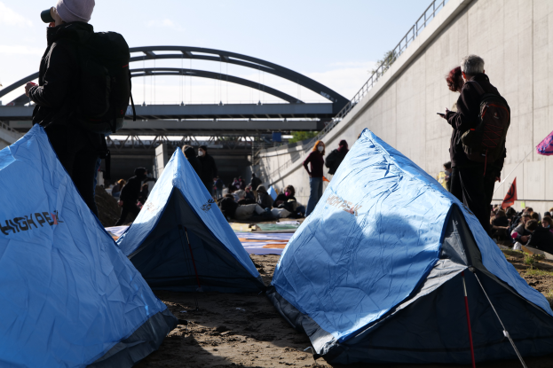 Eine Gruppe von Menschen sitzt auf einem sandigen Strand neben Zelten, mit einer Wand auf der rechten Seite und einer Brücke im Hintergrund, unter einem bewölkten Himmel, tragend Taschen und haltend Gegenstände, was auf eine Klimaprotest hingedeutet.