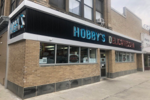 Außenansicht von Hobby's Delicatessen in Chicago, Illinois, mit Glasfenstern und -türen und einer Namensschildtafel, einem Laternenmast davor und einem bewölkten Himmel im Hintergrund.