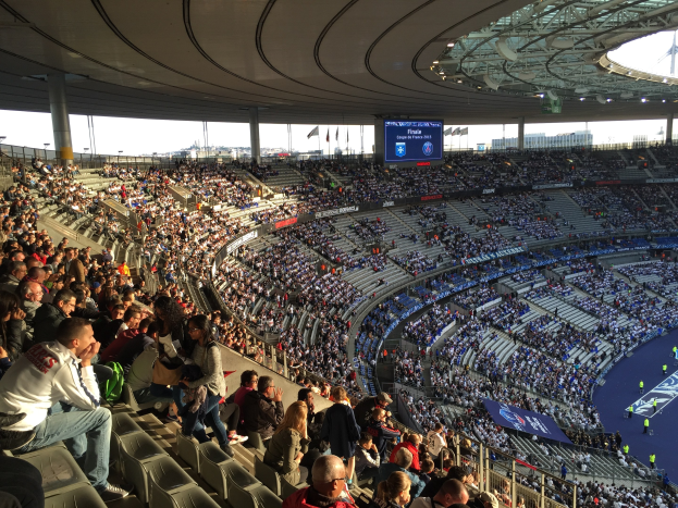 Große Zuschauermenge in einem Stadion bei einem Fußballspiel mit einer Bühne, Fahnen, Stangen, einem Bildschirm und dem Allianz Stadion in München, Deutschland im Hintergrund.
