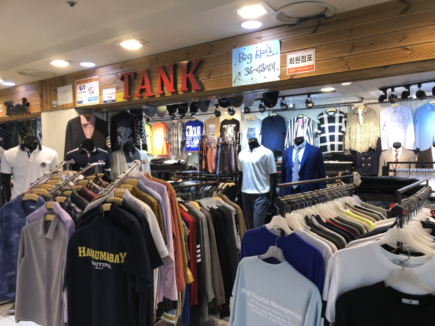 Ein modernes Bekleidungsgeschäft mit zahlreichen Shirts auf Ständern und Schaufensterpuppen in aktuellen Outfits, mit beleuchteter Decke und informativen Wandtafeln.