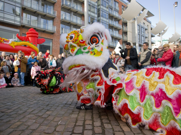 Ein lebendiges chinesisches Neujahrsfest in Amsterdam mit einer Löwen-Tanz-Performance und einer Zuschauermenge, darunter einige, die das Ereignis fotografieren, vor einer Kulisse aus Gebäuden, Laternenmasten und einem klaren blauen Himmel.