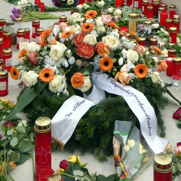 Ein Mahnmal in Berlin, Deutschland, mit einem Strauß Blumen und Kerzen, die vor einer Wand auf dem Boden plaziert sind, umgeben von Blättern und anderen Gegenständen.