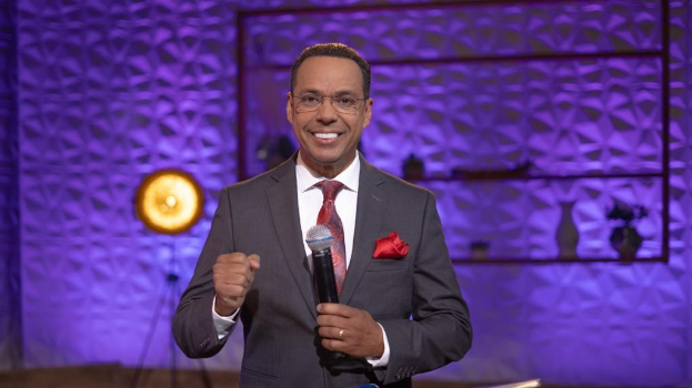 Stephen Colbert, Moderator von "The Daily Show mit Trevor Noah", steht auf einer Bühne und hält ein Mikrofon in der Hand, trägt einen Anzug, Krawatte und eine Brille, während im Hintergrund ein Scheinwerfer und ein Ständer zu sehen sind.