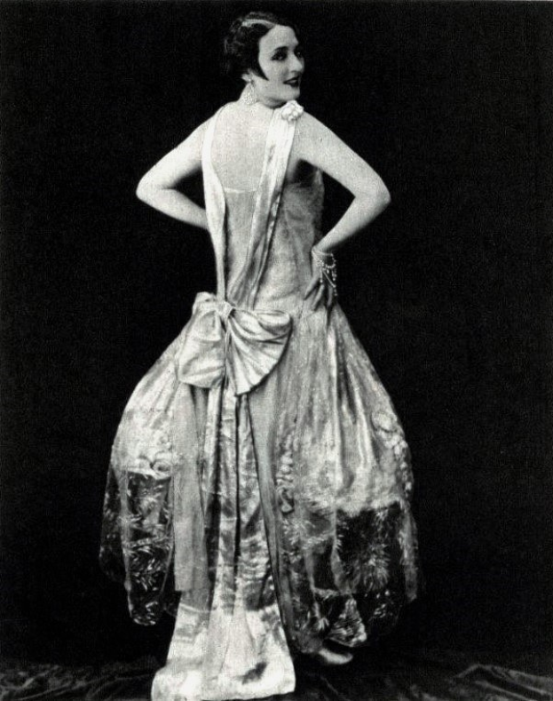 Eine Frau in einem Kleid im Stil der 1920er Jahre mit hohem Kragen, langen Ärmeln und einem ausladenden Rock steht selbstbewusst mit einer eleganten Hochsteckfrisur und dezentem Make-up.