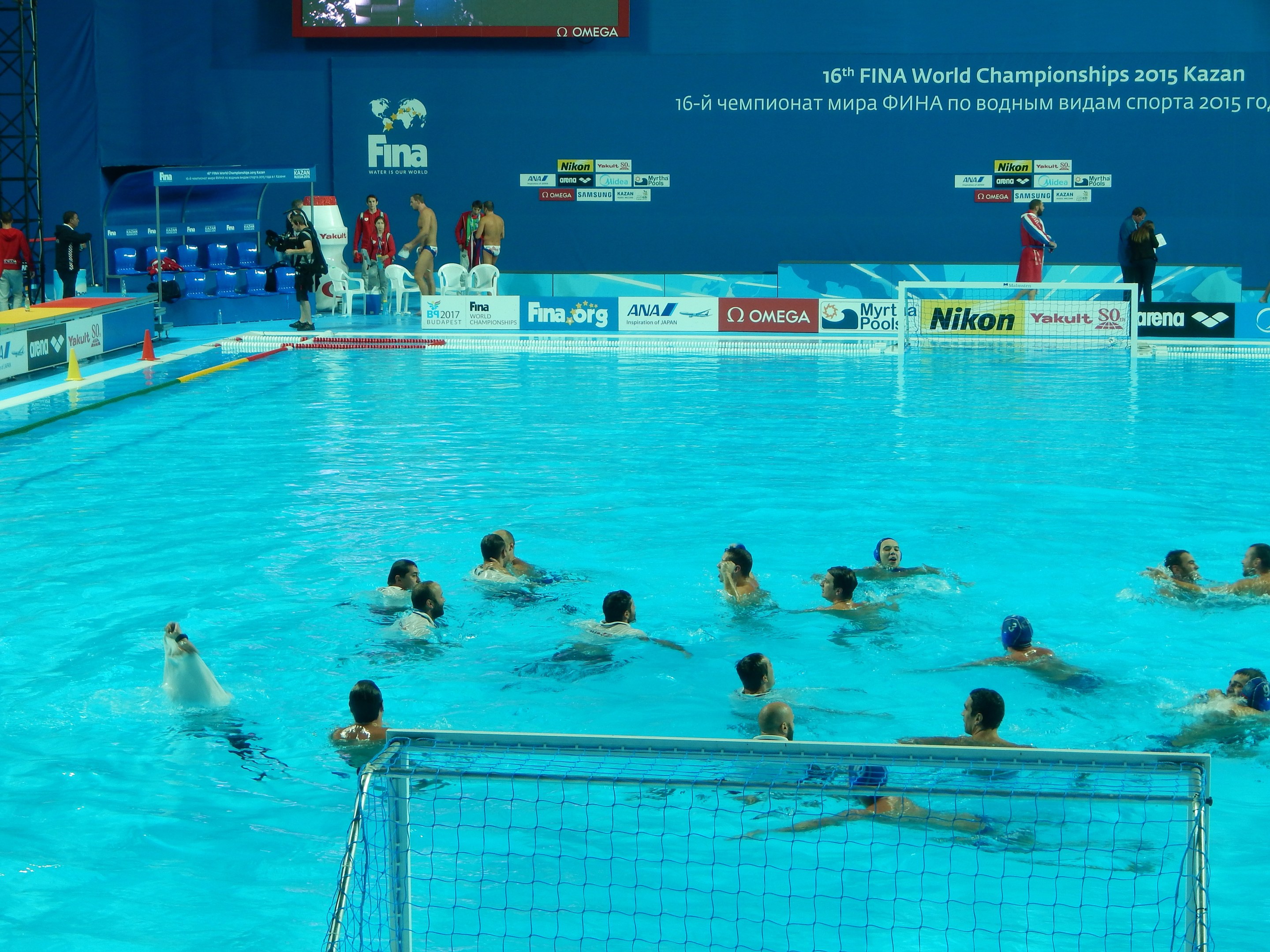 Water polo players competing in a pool during the FINA World Championships 2015 Kazan, with spectators and scoreboards in the background.
