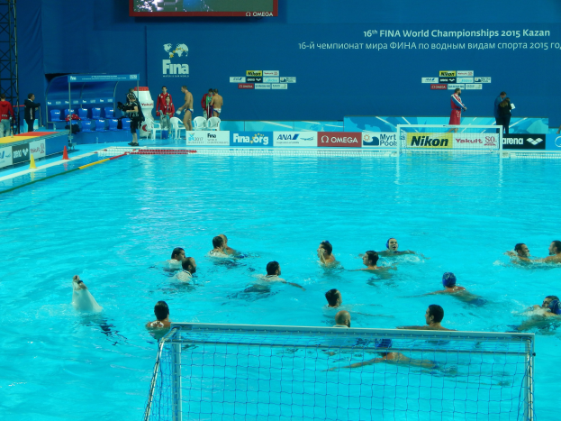 Water polo players competing in a pool during the FINA World Championships 2015 Kazan, with spectators and scoreboards in the background.