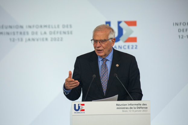 Ein Mann im Anzug und Krawatte hält eine Rede an einem Podium mit zwei Mikrofonen und einem Papier, vor einem Banner mit der Aufschrift "Ministres de la Defense".