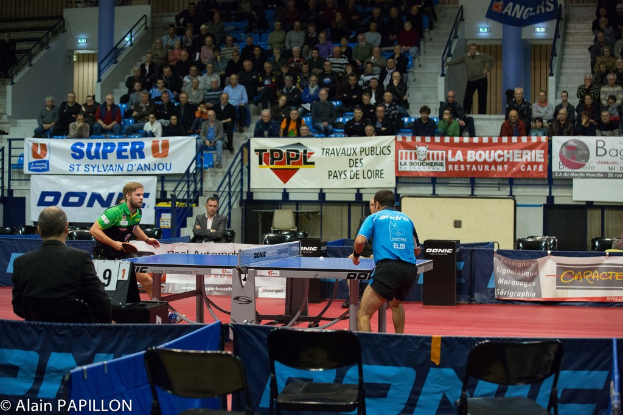 Zwei Männer spielen Tischtennis drinnen, mit mehreren Zuschauern, die stehen und sitzen, einigen leeren Stühlen und Werbeplakaten im Hintergrund.