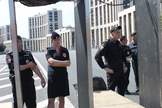 Eine Gruppe von Polizisten in Uniform auf einer Straße mit Masten und Drähten im Vordergrund, Gebäuden und Bäumen im Hintergrund unter einem klaren blauen Himmel.