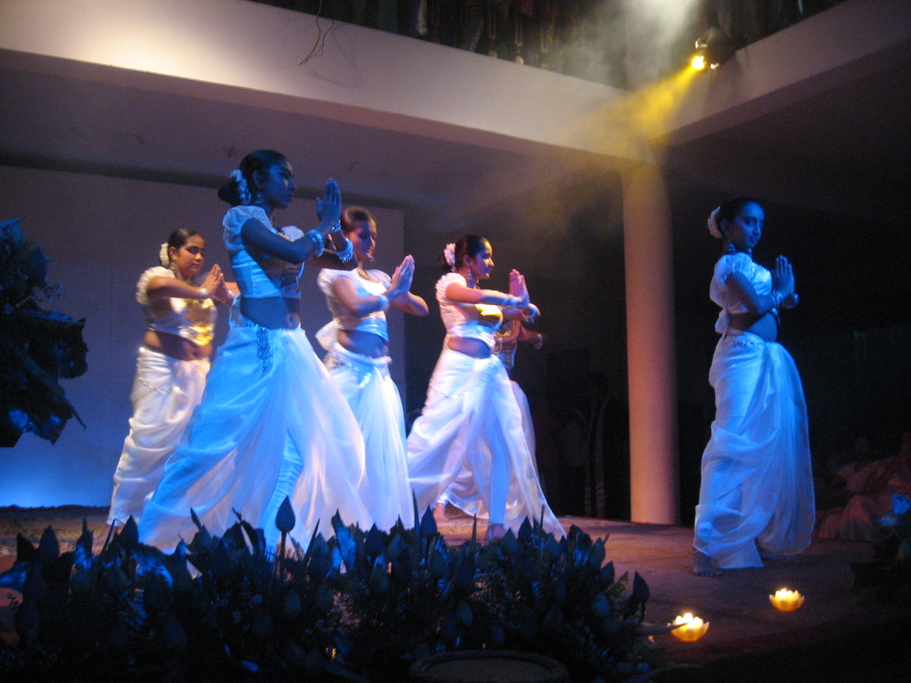 Eine Gruppe von Menschen tanzt auf einer Bühne mit Pflanzen davor und einer Gruppe von Menschen, die zuschauen.