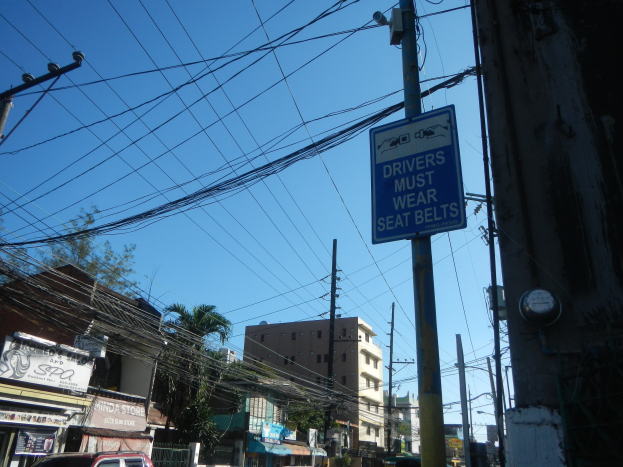 Eine Stadtstraße mit Autos, Strommästen mit Kabeln, Gebäuden, Bäumen und Schildern, auf denen ein "Fahrer müssen den Sicherheitsgurt tragen"-Schild zu sehen ist.