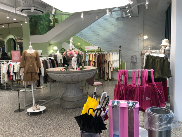 Das Innere eines Bekleidungsgeschäfts mit Schaufensterpuppen, hängenden Kleidern, einem zentralen Brunnen, einem Blumenstrauß in einer Vase, einem Müllbehälter, Taschen und Deckenleuchten.