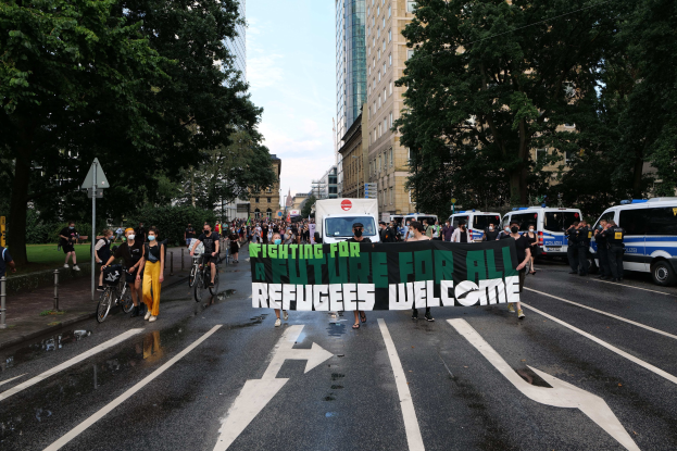 Eine Gruppe von Menschen, die eine Straße entlanggehen und ein Banner mit der Aufschrift "Flüchtlinge Willkommen" halten, mit parkenden Fahrzeugen, Radfahrern und Gebäuden an der Straße.