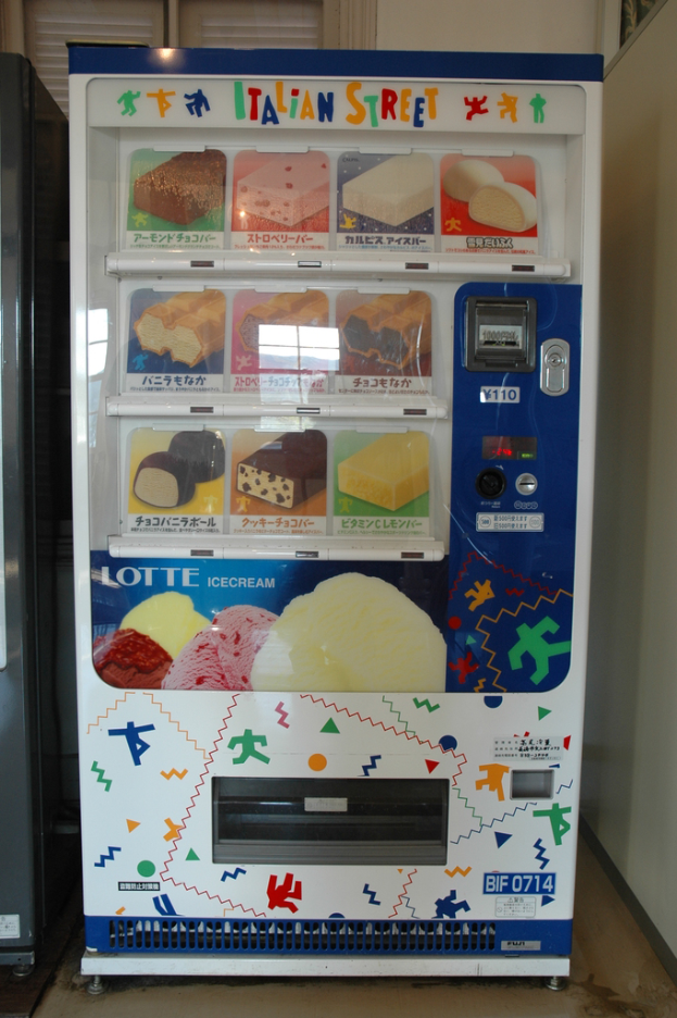 Ein Snackautomat mit Snacks, einer Münzeinwurföffnung oben und einem Spender unten, der an einer Wand mit einem Fenster im Hintergrund steht.