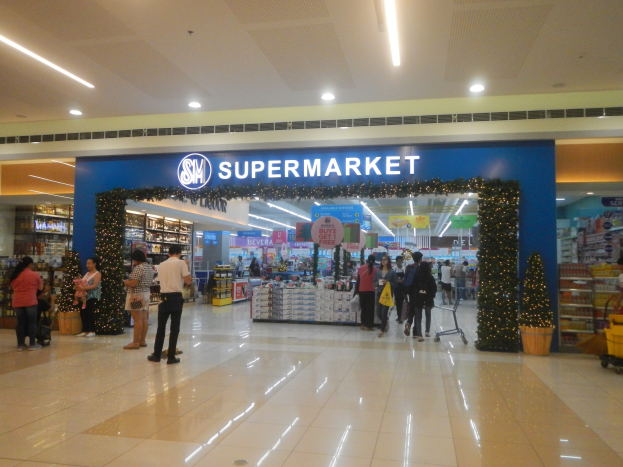 Eine Gruppe von Menschen, die vor einem Supermarkt stehen, einige halten Einkaufstaschen, mit einem Einkaufswagen, Topfpflanze, Gestellen mit Gegenständen, Schildern und einer Decke mit Deckenleuchten im Hintergrund.