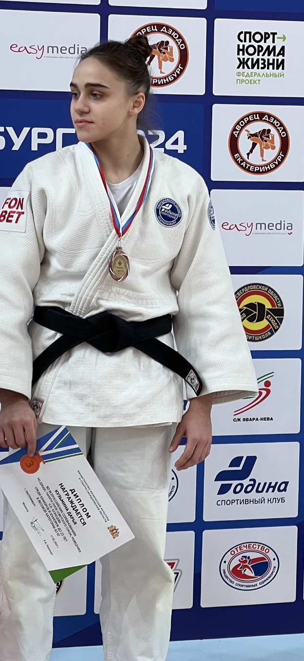 Frau in weißem Judo-Anzug mit schwarzem Gürtel auf einem Podest, die eine Medaille und ein Zertifikat hält, mit einem logo-bedeckten Board im Hintergrund.
