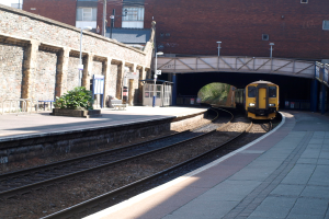 Ein Zug fährt auf den Schienen mit Plattformen auf beiden Seiten, eine Hauswand links, Straßenlaternen, Pflanzen, Bänke und eine Kabine davor, eine Brücke über die Plattformen und Gebäude und Masten im Hintergrund.