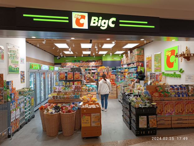 Eine Person in einem weißen und blauen Kleid steht im Vordergrund eines hell erleuchteten Supermarktgangs mit verschiedenen Lebensmitteln, Kühlgeräten, Wandtafeln und Deckenlampen.