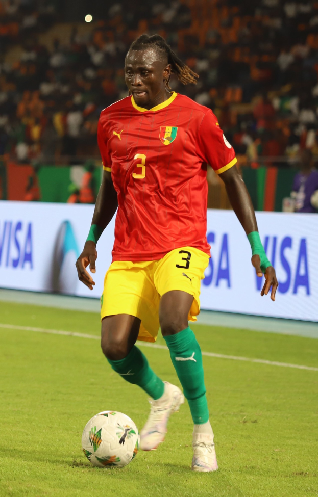 Ein Mann in rot-gelber Uniform spielt Fußball auf einem Feld, umgeben von Zuschauern auf den Tribünen und Werbetafeln im Hintergrund.