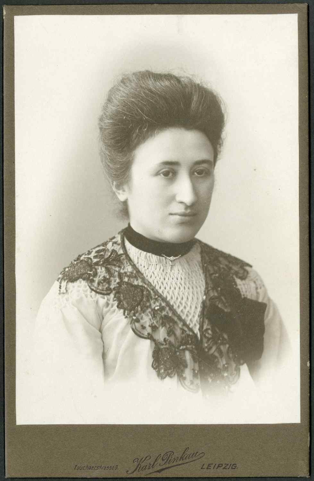 Schwarz-weißes Foto einer Frau in einem weißen Kleid mit einem gemusterten Schultertuch, identifiziert als Karl Pinkau Leipzig, die direkt in die Kamera schaut und einen ernsten Gesichtsausdruck hat.