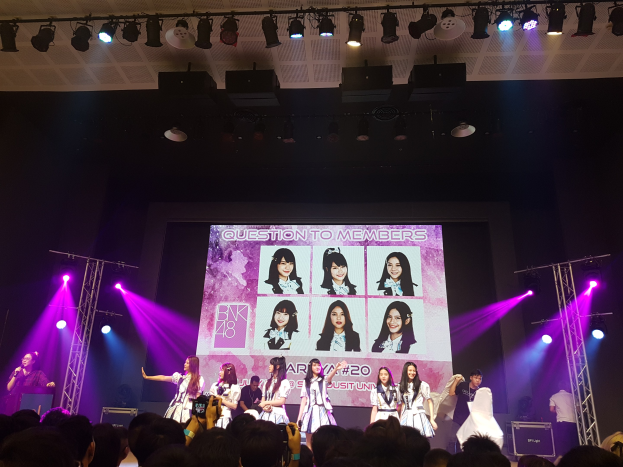 Eine Gruppe von Menschen, die auf einer hell erleuchteten Bühne spielen, mit einer Menge darunter, mit einem Sänger mit Mikrofon und einem Musiker, der ein Instrument spielt, während ein großer Bildschirm Bilder der K-Pop-Gruppe BK48 anzeigt.