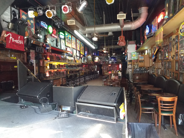 Eine Bar mit Tischen, Stühlen, Sofas, einem Müllcontainer, elektronischen Geräten, Lampen, Texttafeln, Wandrahmen und Flaschenregalen unter einer Decke mit Lampen und einem Rohr, das auf eine Live-Musik-Bar hindeutet.