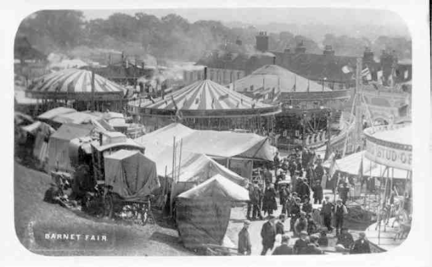 Ein altes Schwarz-Weiß-Foto einer belebten Faschingszene mit Zelten, Menschen, Karren und Gebäuden im Hintergrund, beschriftet mit "Barnet Fair, London, England".