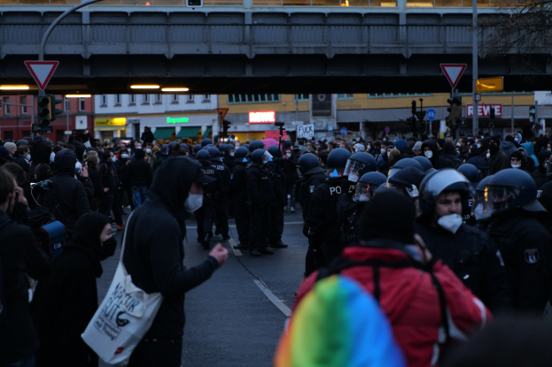 Eine große Gruppe von Menschen vor einer Reihe von Polizisten, einige mit Helmen und Taschen oder Kameras, mit Gebäuden, Schildern, Verkehrszeichen, einer Brücke und einem Baum im Hintergrund.