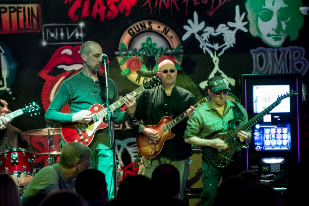 Drei Männer auf einer Bühne mit Gitarren, umgeben von einer Menge und einem Graffiti oder einer Wandmalerei im Hintergrund.